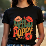 A person wearing a black short-sleeve T-shirt with a large retro-style graphic reading “WILD POPPY” in green, orange, and yellow, surrounded by red-orange poppy flowers. The photo is cropped from the neck to the hips, with a blurred outdoor street background.