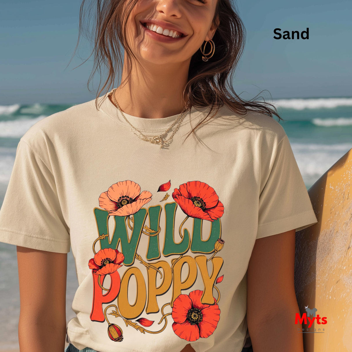 A woman wearing a Sand-colored graphic T-shirt with a colorful 'WILD POPPY' design, standing on a beach next to a surfboard. The text 'Sand' is visible in the top right corner, and a 'Myts' brand logo is in the bottom right corner.