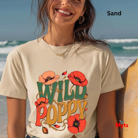 A woman wearing a Sand-colored graphic T-shirt with a colorful 'WILD POPPY' design, standing on a beach next to a surfboard. The text 'Sand' is visible in the top right corner, and a 'Myts' brand logo is in the bottom right corner.