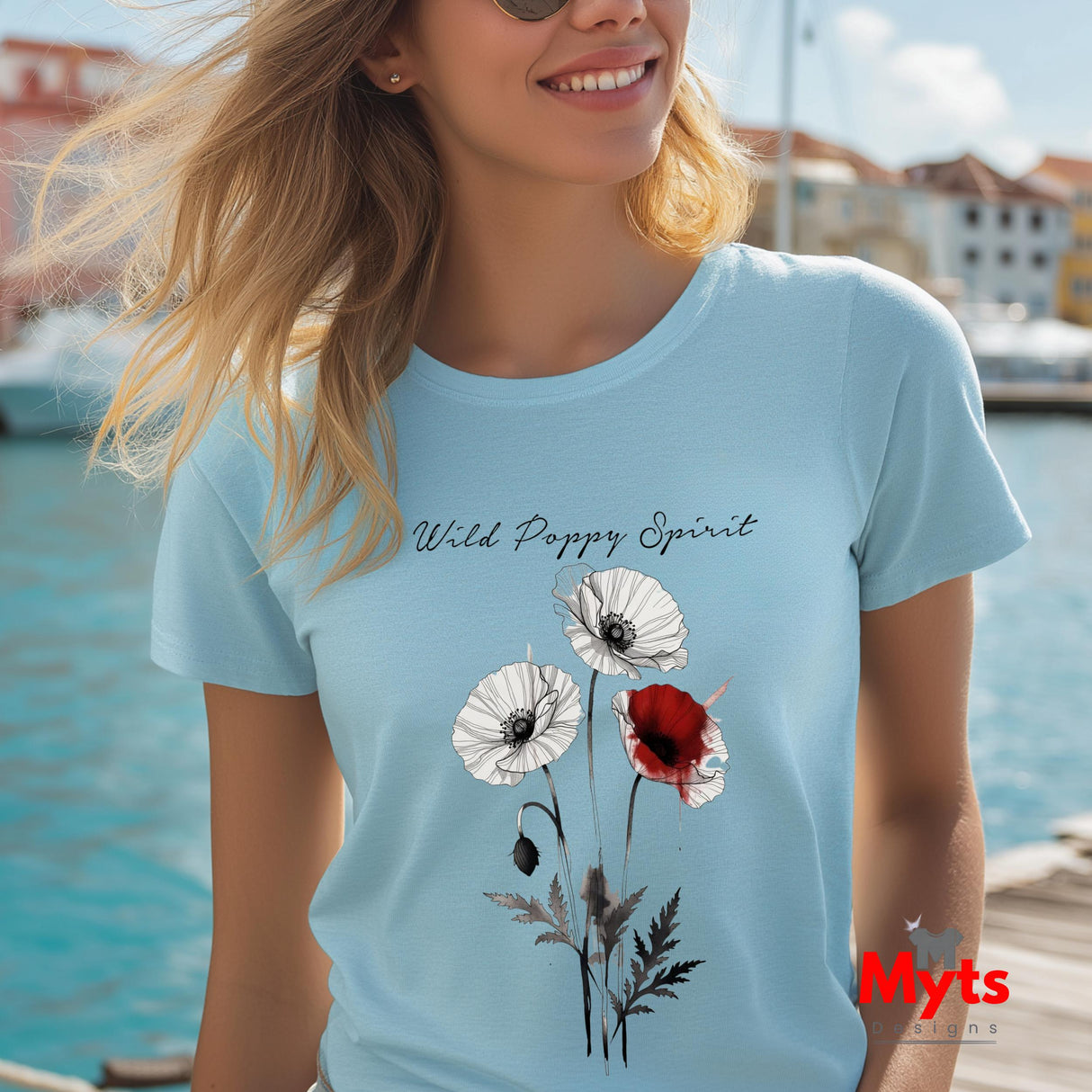 A woman wearing a light blue t-shirt with a 'Wild Poppy Spirit' graphic print featuring white and red poppies, set against a sunny outdoor waterfront background.