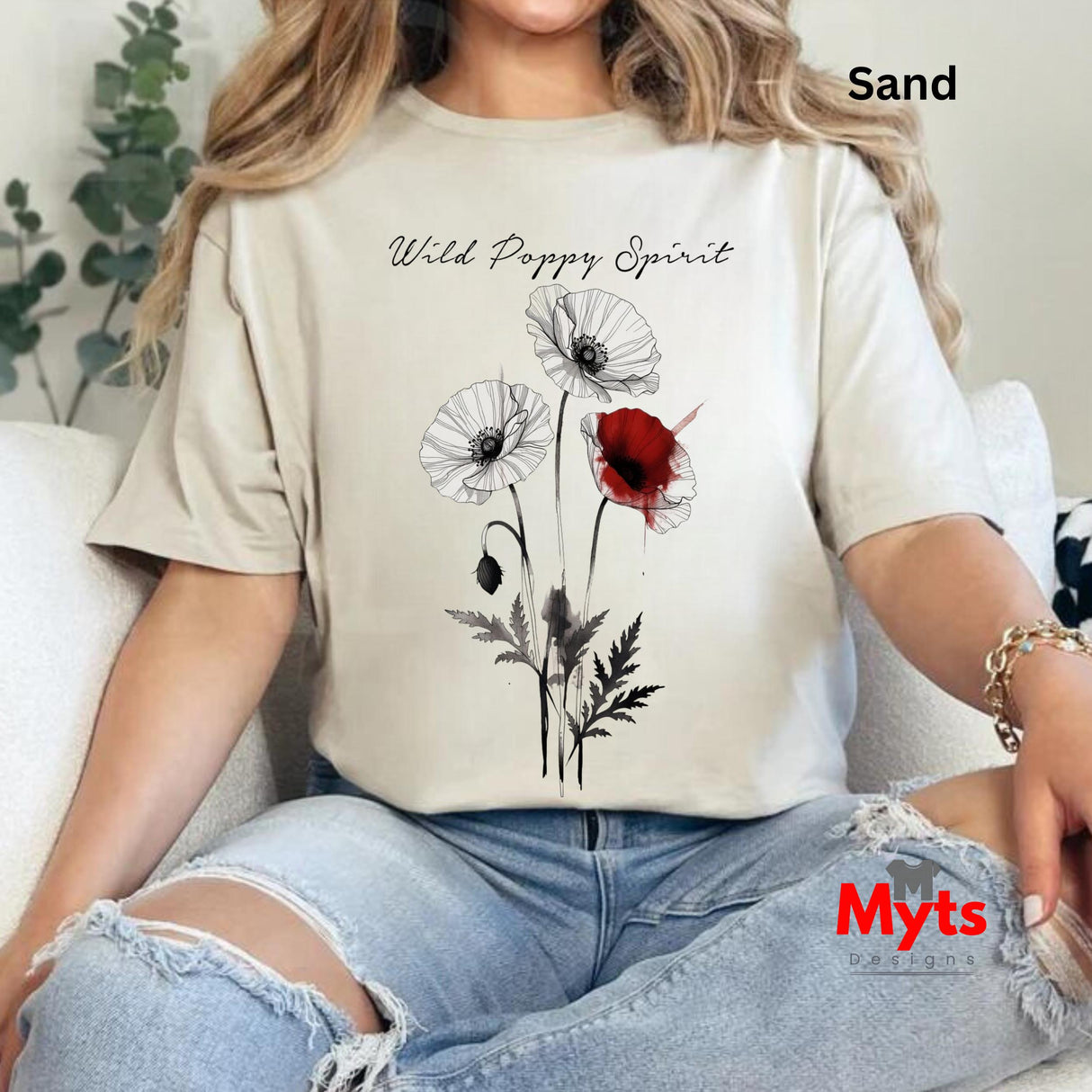A woman wearing a sand-colored t-shirt with a black and red poppy graphic and the text 'Wild Poppy Spirit', paired with distressed light blue jeans, sitting on a white couch.