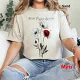 A woman wearing a sand-colored t-shirt with a black and red poppy graphic and the text 'Wild Poppy Spirit', paired with distressed light blue jeans, sitting on a white couch.