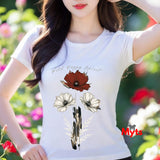 A woman wearing a white short-sleeve t-shirt with a graphic print of wild poppies and the text 'Wild Poppy Spirit'. The background is a blurred garden with green foliage and pink flowers.
