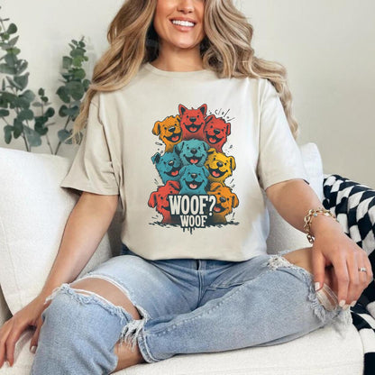 A woman sitting on a comfy couch wearing a Sand T-shirt featuring a vibrant graphic of cartoon puppies stacked in red, orange, blue, and yellow hues, with the playful text “WOOF? WOOF” below the design.