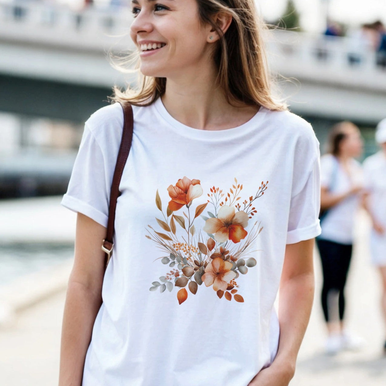 A woman wearing a white t-shirt with a floral design is outdoors by a waterfront.