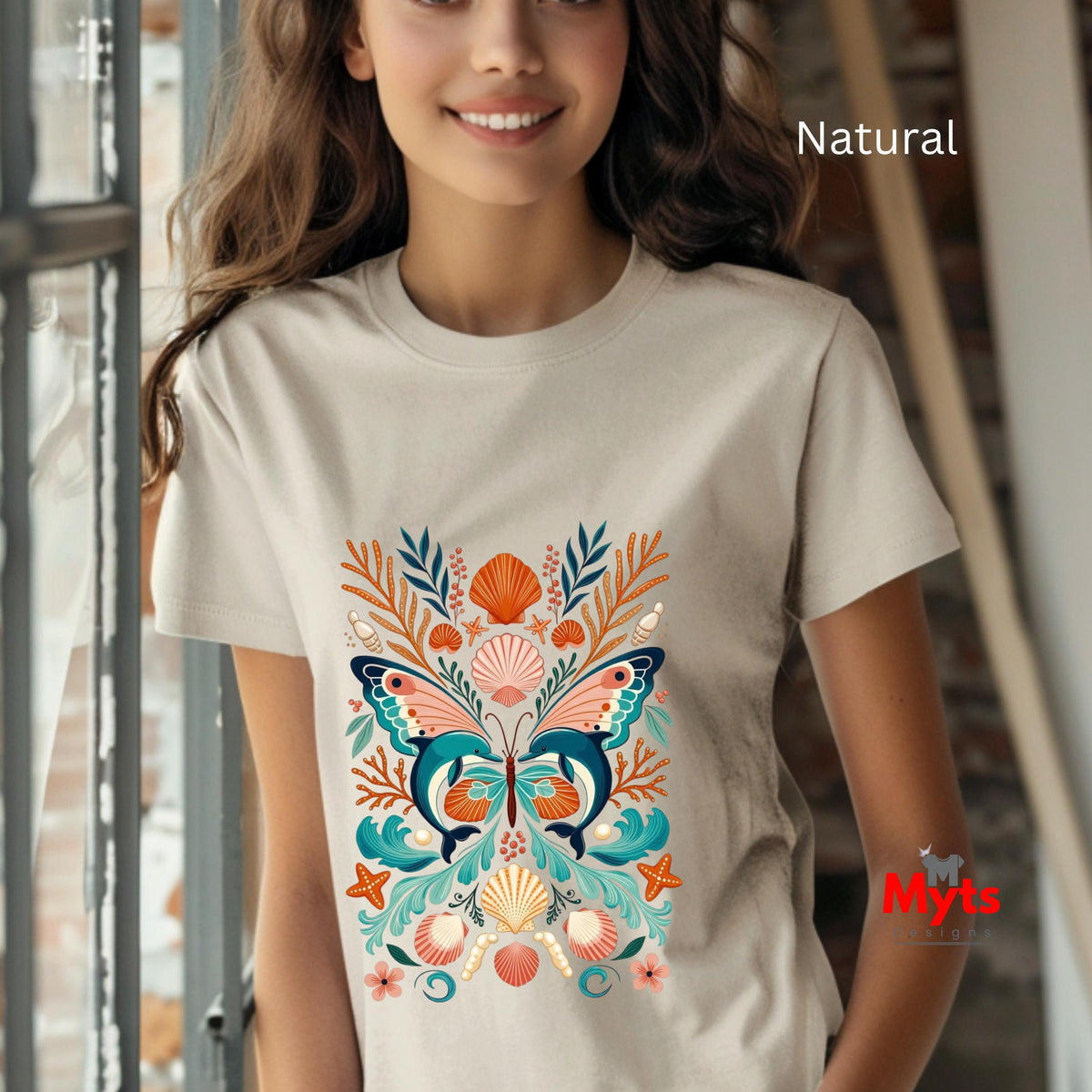 A woman wearing a natural beige t-shirt with a colorful butterfly and seashell graphic print, set against a softly blurred indoor background.