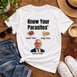 White Know Your Parasites T-shirt displayed flat on a wooden surface with jeans, trainers, hat and coffee.