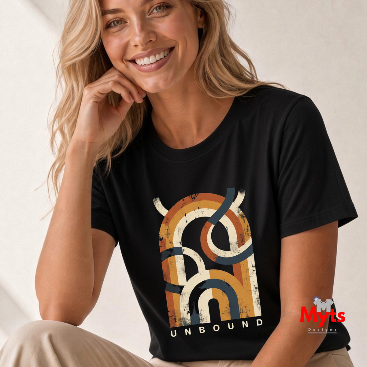 A smiling blonde woman wearing a black short-sleeve graphic T-shirt with a large distressed multicolor abstract design and the word 'UNBOUND' printed below it, paired with light beige pants against a soft neutral background.