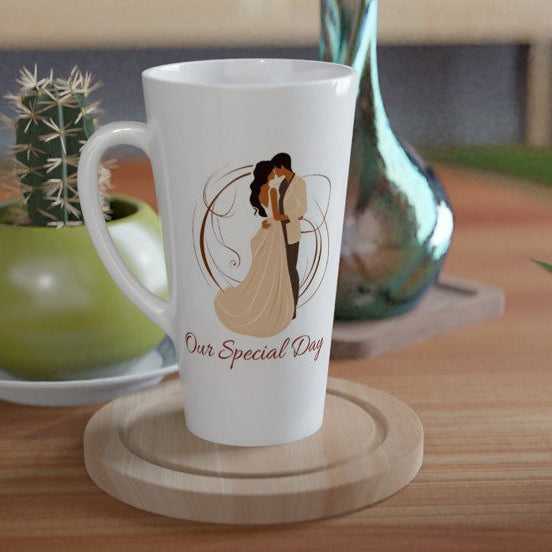 A white ceramic latte mug with a custom illustration of a couple and the text 'Our Special Day' sits on a wooden coaster on a wooden table, styled with a small potted cactus and a green glass vase in the background.
