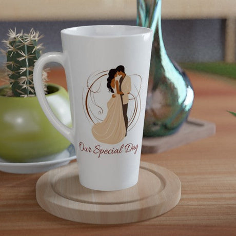 A white ceramic latte mug with a custom illustration of a couple and the text 'Our Special Day' sits on a wooden coaster on a wooden table, styled with a small potted cactus and a green glass vase in the background.