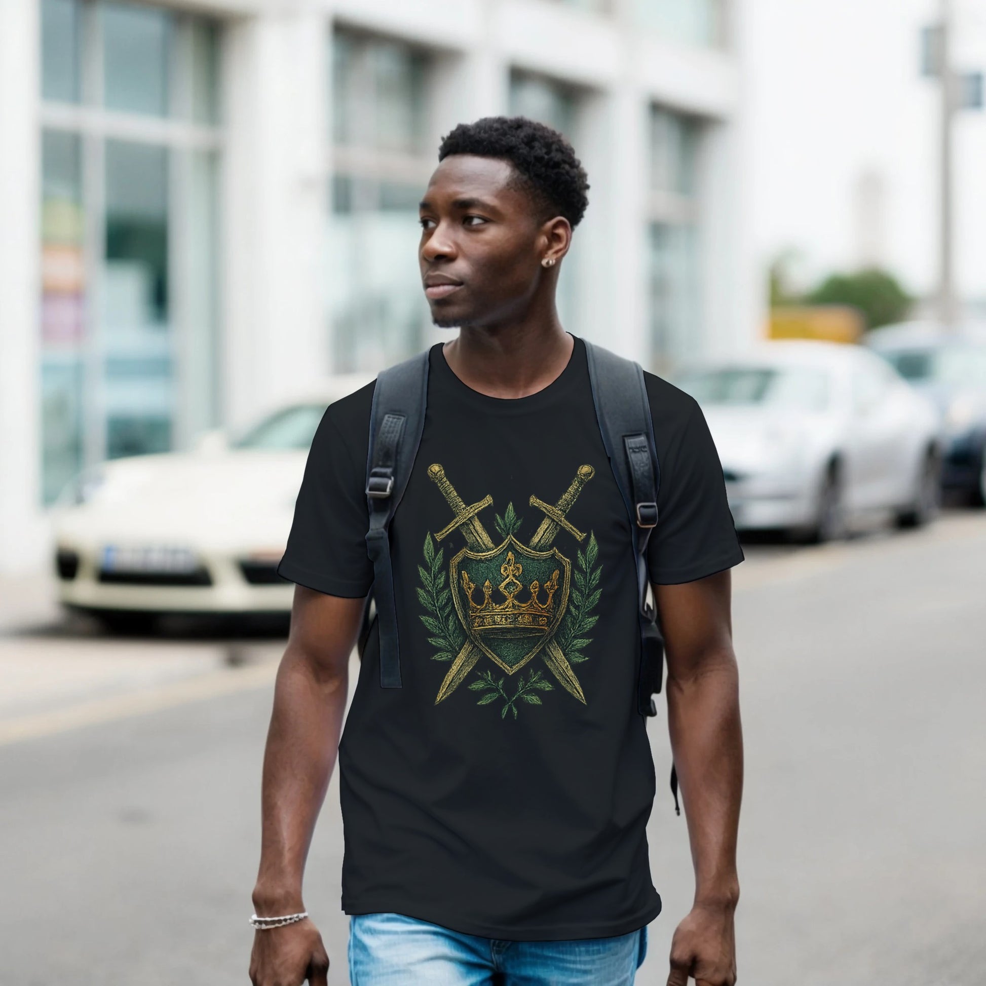 Man wearing a black T-shirt with an embroidered-style design of a green shield featuring a gold crown, flanked by crossed swords and laurel leaves—regal, medieval-inspired graphic centered on the chest.