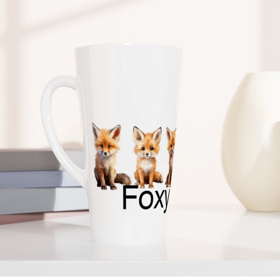 A tall white ceramic mug with a handle, featuring a printed graphic of three red foxes and the partial text 'Foxy' in black. The mug is styled on a white surface next to stacked books, with two minimalist ceramic vases in the background.