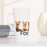 A tall white ceramic mug with a handle, featuring a printed graphic of three red foxes and the partial text 'Foxy' in black. The mug is styled on a white surface next to stacked books, with two minimalist ceramic vases in the background.