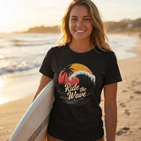 Woman on a beach holding a surfboard wearing a black t-shirt with a wave design and text.