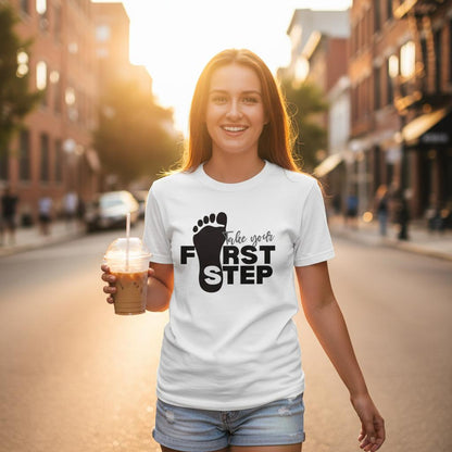 Woman wearing a white t-shirt with 'First Step' design, holding a drink on a city street.