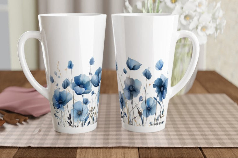 Two tall white ceramic mugs with blue floral illustrations, displayed on a checkered placemat on a wooden table with a vase of flowers in the background.