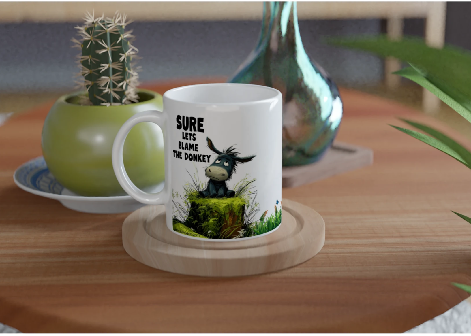 A white ceramic coffee mug sitting on a wooden coaster with a cute donkey sitting on a moss log, with the caption "Sure, Let's Blame the Donkey"