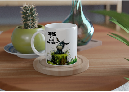 A white ceramic coffee mug sitting on a wooden coaster with a cute donkey sitting on a moss log, with the caption "Sure, Let's Blame the Donkey"