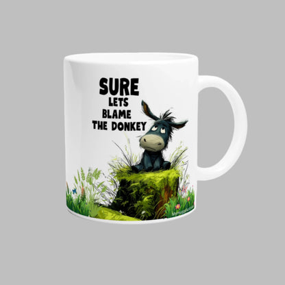 White ceramic coffee mug with a cute donkey sitting on a moss log, with the caption "Sure, Let's Blame The Donkey"