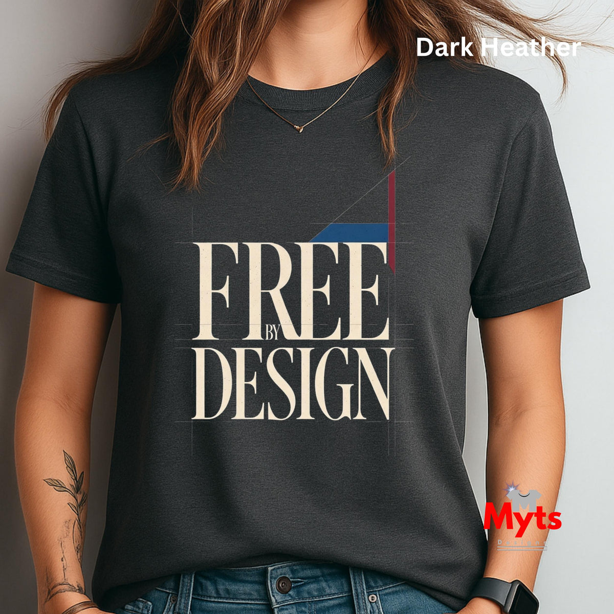 A woman wearing a dark heather charcoal T-shirt with a graphic print that reads 'FREE BY DESIGN' in cream lettering, with a blue and red geometric accent. The image includes 'Dark Heather' text in the top right and a 'Myts' watermark in the bottom right.
