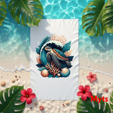 A white beach towel featuring a colorful graphic of a sea turtle, waves, and coral, laid flat on a sandy beach with tropical leaves, flowers, and water in the background.