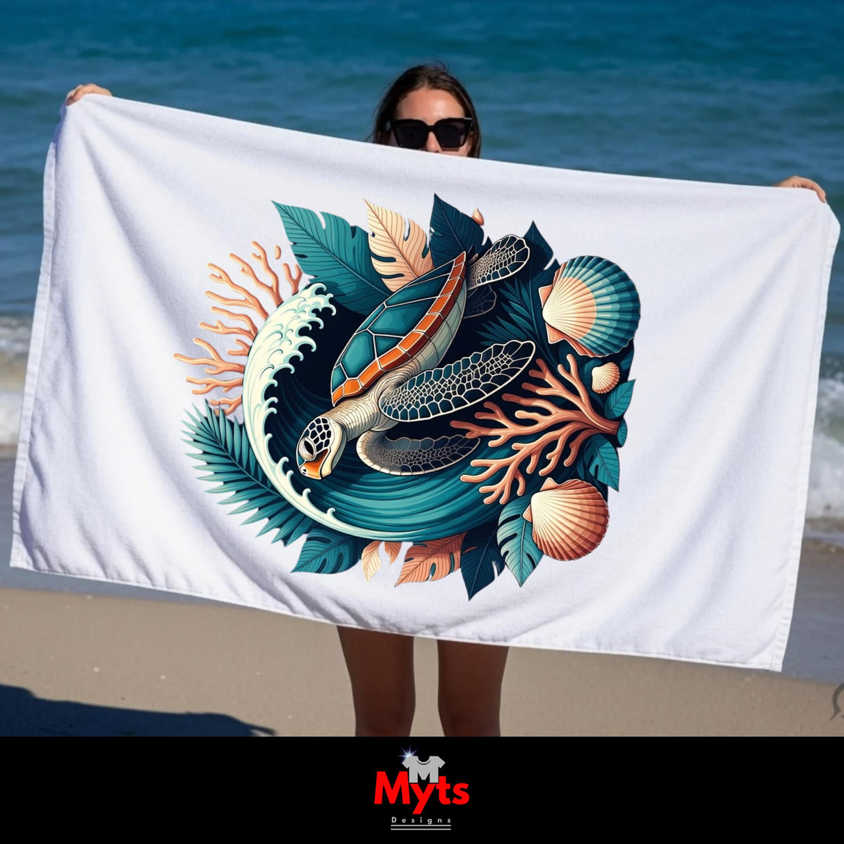 A person on a beach holds up a large white beach towel featuring a colorful graphic of a sea turtle, waves, leaves, and seashells. The background shows the blue ocean and sand.