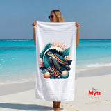 A woman on a sandy beach holds up a large white beach towel featuring a colorful graphic of a sea turtle, coral, and seashells. The background shows a turquoise ocean and blue sky.