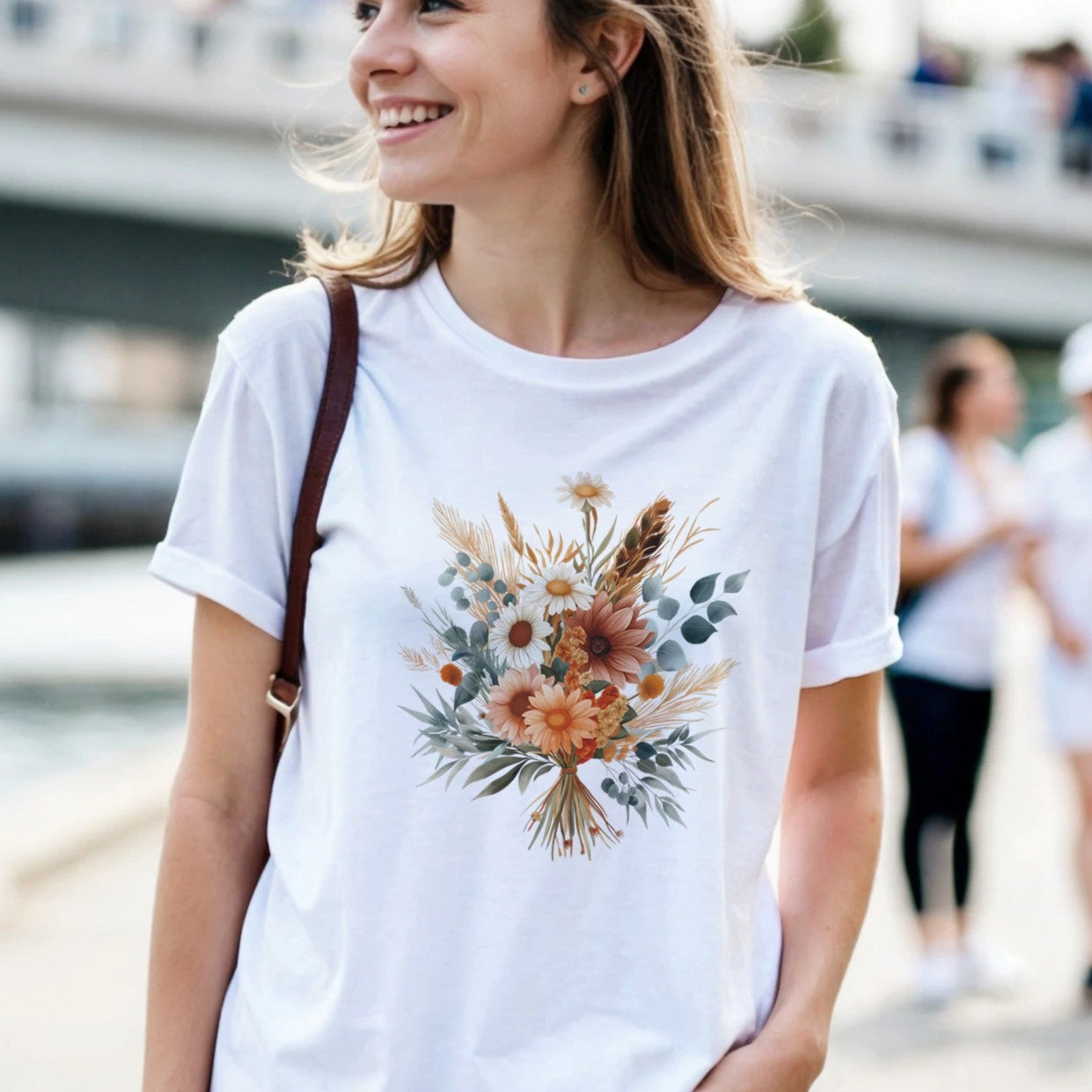 A woman wearing a white t-shirt with a floral design is outdoors by a waterfront.