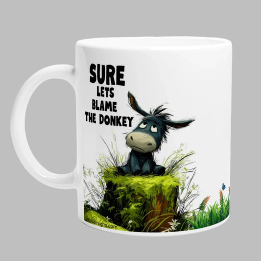 White ceramic coffee mug with a cute donkey sitting on a moss log, with the caption "Sure, Let's Blame The Donkey"