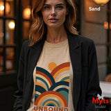 A woman wearing a Sand graphic t-shirt with a retro rainbow design and the word 'UNBOUND' underneath, styled with a black blazer. The image has a warm, blurred indoor background with bokeh lights.