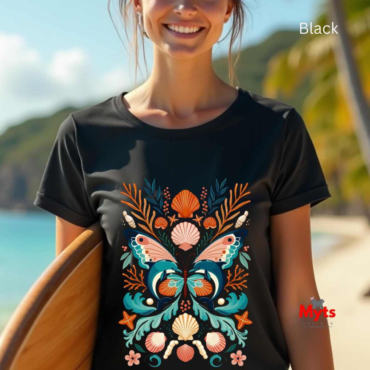 A woman wearing a black short-sleeve t-shirt with a colorful butterfly and seashell graphic, standing on a beach with a surfboard.