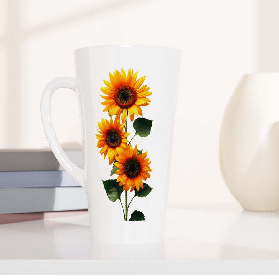 A tall white ceramic latte mug with a handle, decorated with a graphic of three yellow sunflowers with green leaves, set on a white surface next to stacked books and two ceramic vases.