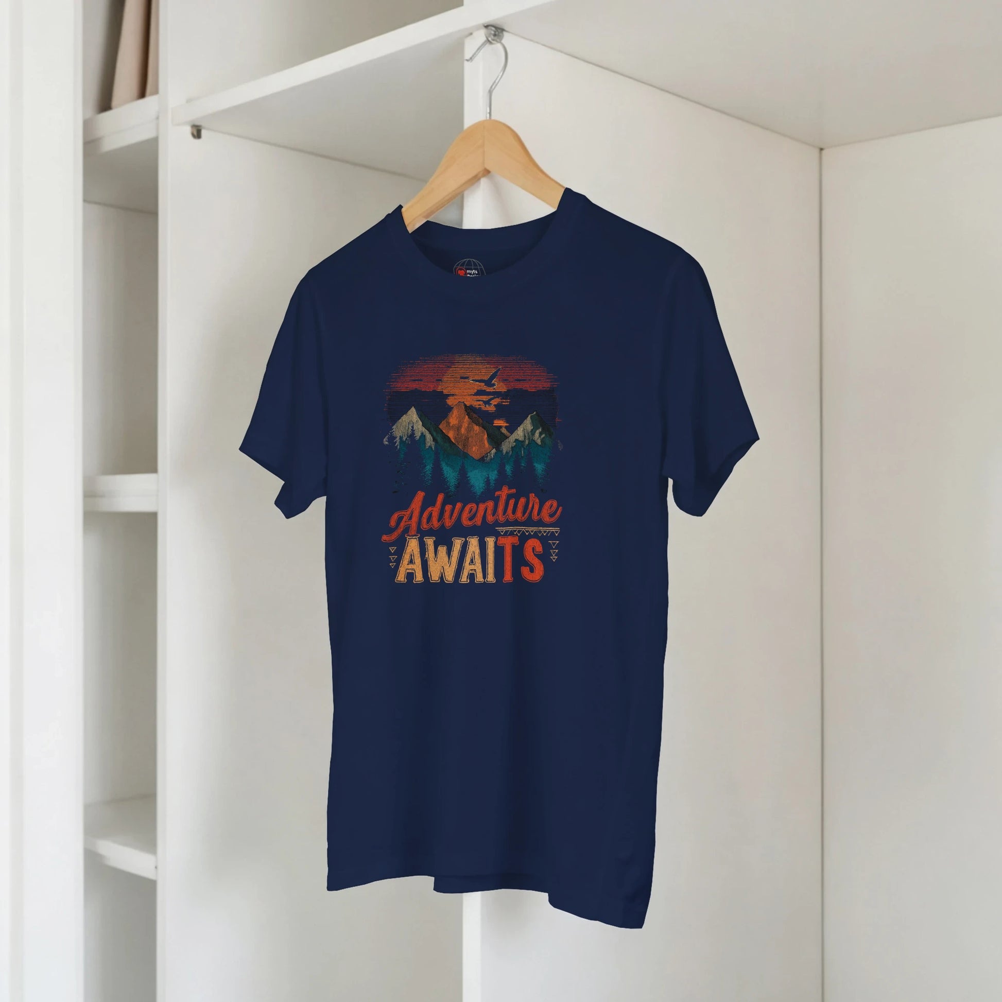 Navy blue t-shirt with 'Adventure Awaits' graphic on a hanger against a white wall.