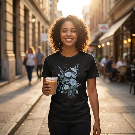 A smiling woman with curly hair stands on a sunlit city street holding an iced coffee. She wears a black short-sleeve T-shirt with a light blue and white floral bouquet graphic on the front.