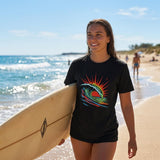 A woman walking on a beach with a surfboard, wearing a black t-shirt with a colorful wave design.