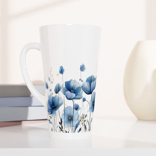A tall white ceramic mug with a blue floral print, styled on a white surface next to stacked books and two neutral-colored ceramic vases.