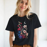 A blonde woman wearing a black short-sleeve t-shirt with a colorful floral and crescent moon graphic, paired with brown pants, standing in a bright indoor setting.