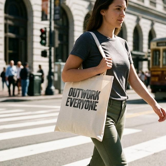 Woman walking on a city street with a tote bag that says 'OUTWORK EVERYONE'.