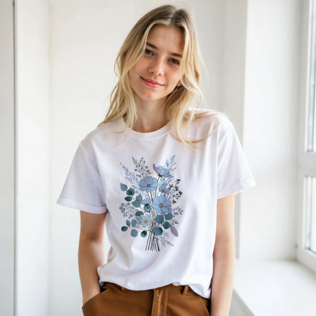 A blonde woman wearing a white short-sleeve crewneck t-shirt with a blue and gray floral bouquet graphic on the front, paired with brown pants, standing in a bright, minimalist room.
