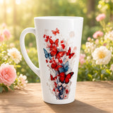 A tall white ceramic mug with a handle, decorated with a vibrant watercolor-style print of red and blue butterflies, placed on a wooden table with a blurred garden background of pink and white flowers.