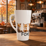 A tall white ceramic latte mug with a handle, featuring a printed graphic of three cute foxes and the partial text 'Foxy' in black, set on a wooden table in a warm, blurred coffee shop background.