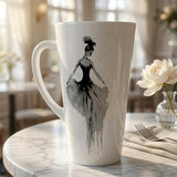A tall white ceramic latte mug with a black and gray watercolor-style illustration of a ballerina, set on a marble table in a softly blurred cafe interior.
