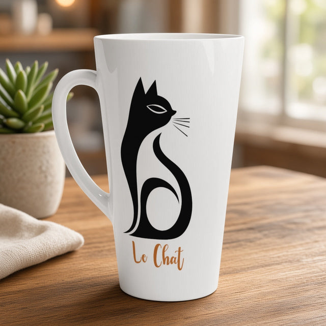 A tall white ceramic coffee mug with a black cat silhouette and the French text 'Le Chat' in brown, displayed on a wooden table in a warm, softly blurred indoor setting.