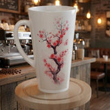 A tall white ceramic latte mug with a pink and red cherry blossom tree design, displayed on a wooden stand in a cozy, blurred coffee shop setting.