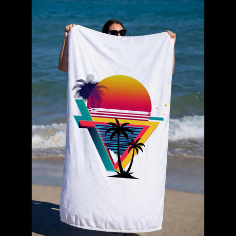 A person on a beach holds up a large white beach towel featuring a colorful retro sunset and palm tree graphic.