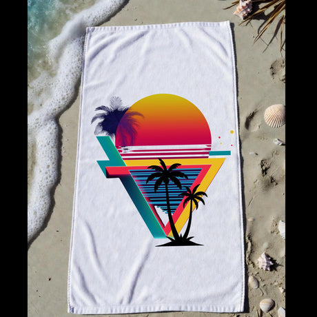 A white beach towel laid flat on sandy beach ground with ocean foam and shells nearby. The towel features a colorful retro sunset and palm tree graphic with overlapping geometric shapes.