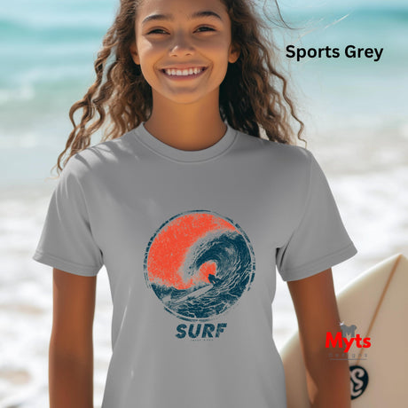 A smiling young woman on a beach wearing a light gray t-shirt with a distressed graphic of a blue and orange surf wave and the word 'SURF' underneath. The text 'Sports Grey' is visible in the top right, and a red 'Myts' brand logo is in the bottom right.