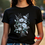 A woman wearing a black short-sleeve crewneck t-shirt with a watercolor-style floral bouquet graphic on the front, featuring white flowers and muted green leaves. The background is a softly blurred outdoor street scene.