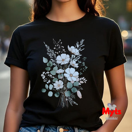 A woman wearing a black short-sleeve crewneck t-shirt with a watercolor-style floral bouquet graphic on the front, featuring white flowers and muted green leaves. The background is a softly blurred outdoor street scene.